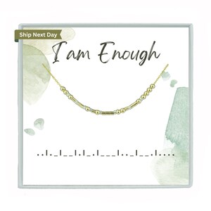 May include: A white box with a green watercolor background. The box contains a gold and silver beaded necklace with the text "I am Enough" written in black cursive.