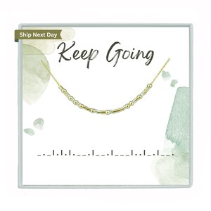 May include: A white box with a green watercolour background. The box has the text "Keep Going" in black lettering and a gold chain with alternating gold and silver beads. The text "Ship Next Day" is in a brown rectangle at the top left corner of the box.