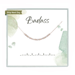 Peut inclure: Un collier délicat en chaîne dorée avec de petites perles dorées et or rose. Le collier est présenté dans une boîte blanche avec un fond aquarelle vert et le mot "Badass" écrit en cursive noire.