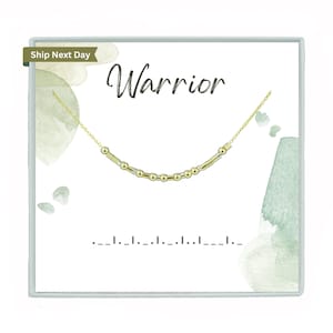 May include: A gold-colored necklace with a Morse code design, presented on a white card with the word "Warrior" in script. The card has watercolor-style green accents and a light blue border.