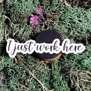 May include: A round, black magnet with the text "I just work here" in white script. The magnet is set on a bed of green moss with small purple flowers.