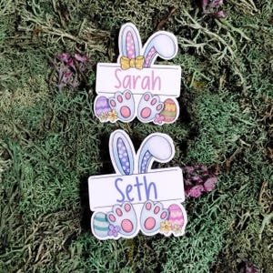 May include: Two Easter-themed name tags with bunny ears, paw prints, and Easter eggs. The top tag says "Sarah" in pink, and the bottom tag says "Seth" in blue. The tags are set against a mossy green background.