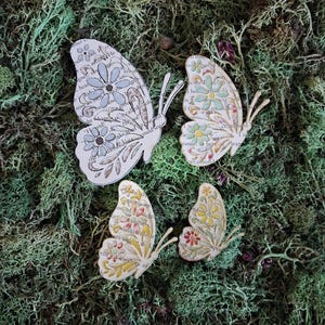 Puede incluir: Cuatro recortes con forma de mariposa hechos de páginas de libro impresas con texto en tinta negra. Cada mariposa presenta patrones florales pintados a mano en tonos azul, verde, amarillo y rojo, con antenas delicadas en las alas.