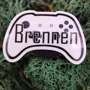 May include: A white controller-shaped sticker with the name "Brennen" in black block letters. The sticker has a black outline and features black button details. The background is a bed of green moss.