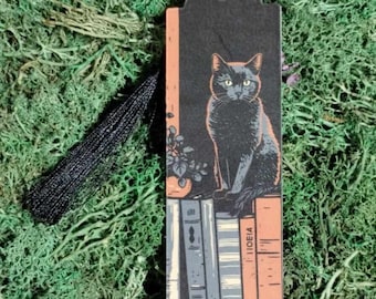 May include: Black cat bookmark with a black tassel. The cat is sitting on a stack of books with the word "IDEA" visible on one of the books.