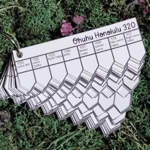May include: A colour swatch book with the text "Ohuhu Honolulu 320" and colour names like "Lemon Mist" and "Summer Lemon." The book is on a metal ring and features hexagonal colour samples. The background is green moss with small purple flowers.
