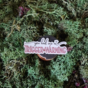 Puede incluir: Una pegatina con el texto "you had me at TRIGGER WARNING" en letras blancas y rosas, sobre un fondo negro. La pegatina está colocada sobre un objeto pequeño, redondo y marrón, rodeado de musgo verde.