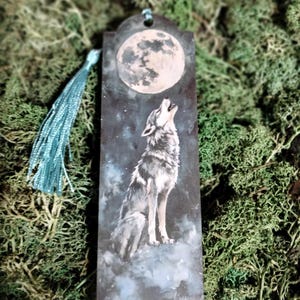 Könnte beinhalten: Ein Holz-Lesezeichen mit einem blauen Quasten-Anhänger zeigt einen Wolf, der zu einem Vollmond heult, vor einem dunkelblauen Sternenhimmel.