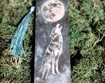 Puede incluir: Un marcapáginas de madera con un borla azul presenta un lobo aullando a la luna llena contra un cielo estrellado azul oscuro.