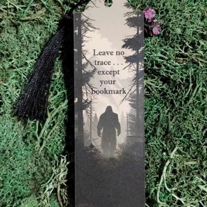 May include: A black and gray bookmark with a silhouette of Bigfoot walking through a forest. The bookmark has the text "Leave no trace... except your bookmark".