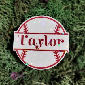 May include: A white baseball-themed sticker with a red border and the name "Taylor" in red letters. The sticker has a glittery finish.