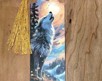 Puede incluir: Un marcapáginas de madera con un borla dorada presenta un lobo aullando a la luna. El lobo está pintado en tonos de gris y blanco sobre un fondo de puesta de sol colorido.