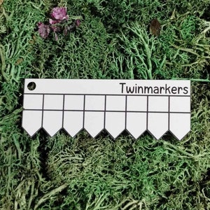 以下が含まれることがあります： 「Twinmarkers」と印刷された、波状の縁を持つ白いプラスチック定規。定規には15個の正方形があります。