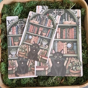 May include: Three rectangular bookmarks featuring a detailed illustration of a black cat sitting in front of a green arched bookshelf filled with various books, potted plants, a small skull, and a globe, all set against a background of green moss.