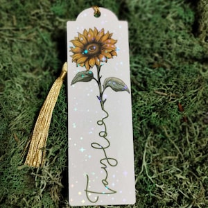Pu&ograve; includere: Un segnalibro bianco con un nappa dorata presenta un girasole giallo con una finitura scintillante e iridescente. La parola "Viaggio" &egrave; scritta in corsivo sul segnalibro.