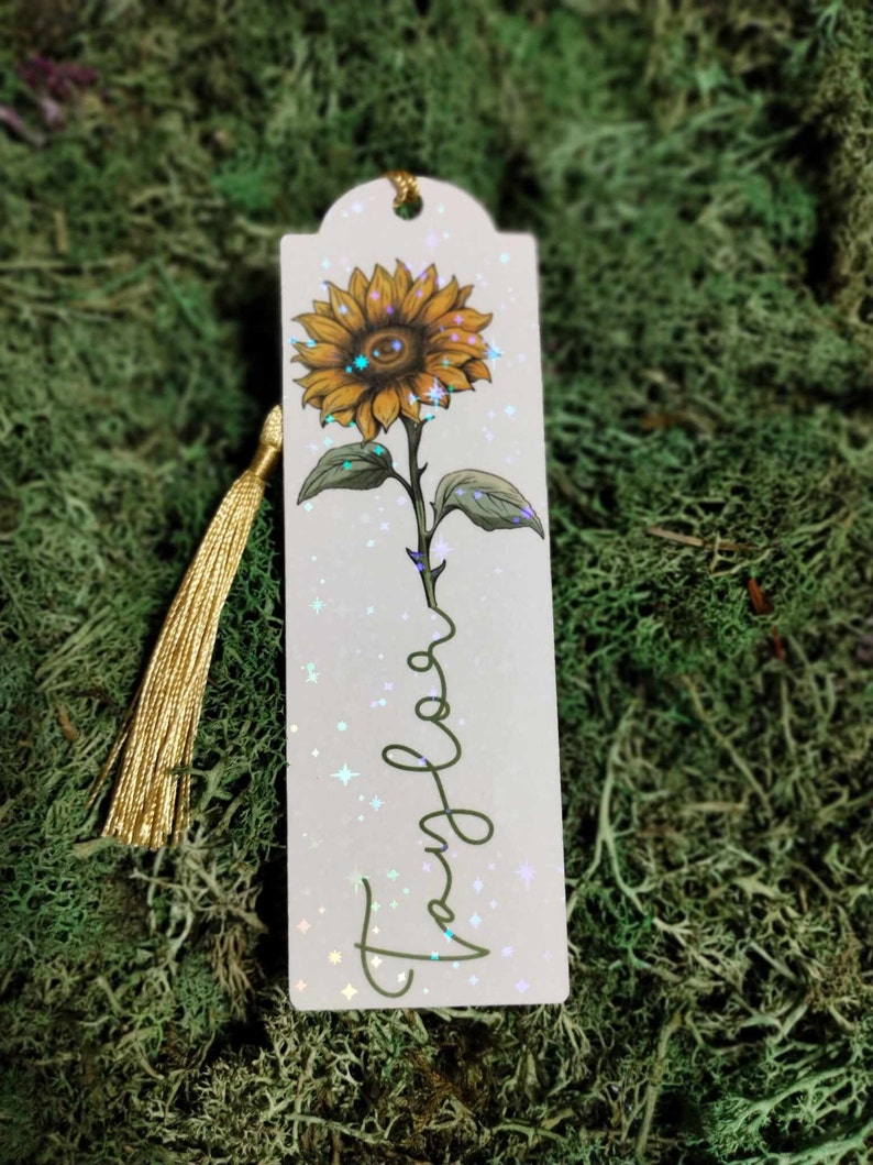 Pu&ograve; includere: Un segnalibro bianco con un nappa dorata presenta un girasole giallo con una finitura scintillante e iridescente. La parola "Viaggio" &egrave; scritta in corsivo sul segnalibro.