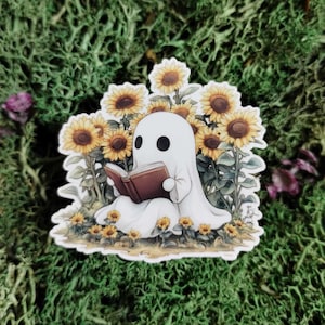 May include: A white ghost sticker reading a brown book, surrounded by sunflowers. The ghost has two black eyes and is draped in a white sheet. The sunflowers are yellow and brown, with green leaves. The sticker is on a green mossy background.