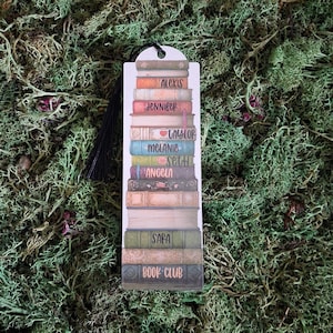 May include: A rectangular bookmark featuring a vertical stack of illustrated books in various colours including tan, pink, blue, green, and brown. The spines are printed with names like Alexis, Jennifer, Taylor, Melanie, Seth, Angela, Sara, and Book Club.