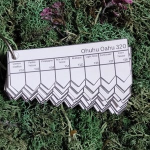 May include: A color swatch card labeled "Ohuhu Oahu 320" with various yellow hues, including "Lemon Chiffon," "Mustard," and "Sunflower." The card is attached to a metal ring and rests on a bed of green moss.