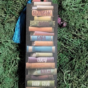 May include: A black bookmark with a stack of colorful books, each with a name on the spine. The names are Elizabeth, Alexis, Sara, Lucy, Faith, and Book Club. The bookmark has a blue tassel.