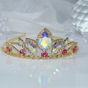 May include: A gold tiara with iridescent and pink rhinestones. The tiara has a delicate design with a pointed center and curved sides.
