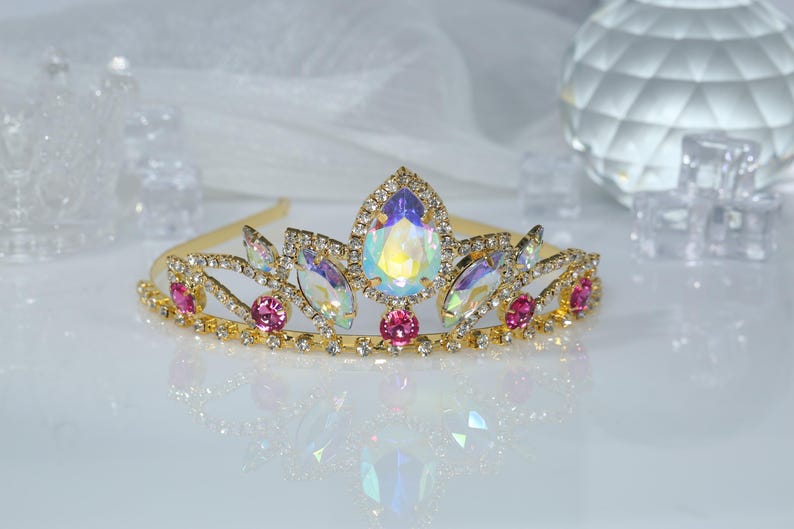 May include: A gold tiara with iridescent and pink rhinestones. The tiara has a delicate design with a pointed center and curved sides.