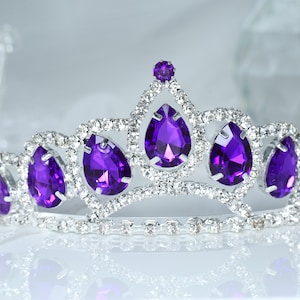 Peut inclure: Une tiare argentée avec une rangée de strass en forme de goutte d'eau violette.