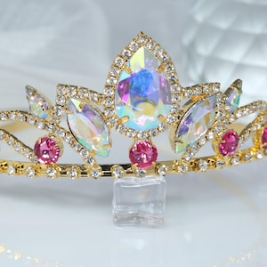 May include: A gold tiara with iridescent and pink rhinestones. The tiara has a pointed crown and is decorated with small, clear rhinestones.