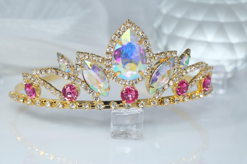 May include: A gold tiara with iridescent and pink rhinestones. The tiara has a pointed crown and is decorated with small, clear rhinestones.