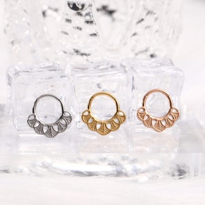 Puede incluir: Tres anillos de septum en plata, oro y oro rosa con un diseño decorativo en forma de abanico. Los anillos se muestran sobre una superficie transparente.