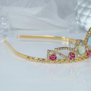May include: A gold tiara with pink and iridescent stones. The tiara has a delicate design with a curved shape and a band that goes around the head.