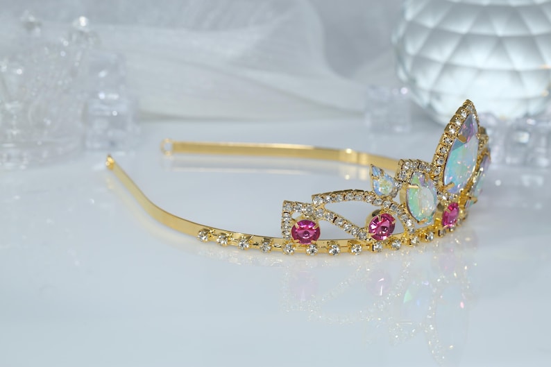 May include: A gold tiara with pink and iridescent stones. The tiara has a delicate design with a curved shape and a band that goes around the head.