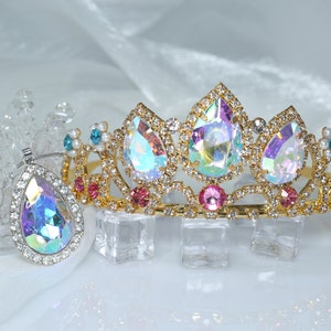 Könnte beinhalten: Eine goldene Tiara mit schillernden Edelsteinen und rosafarbenen Akzenten. Die Tiara liegt auf einer weißen Oberfläche mit Eiswürfeln.