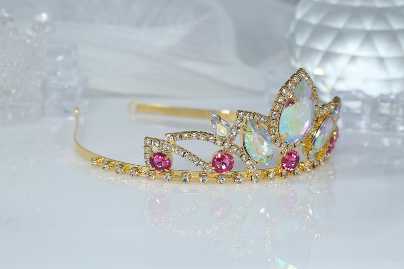 May include: A gold tiara with pink and iridescent stones. The tiara has a delicate design with a pointed crown and a band that wraps around the head.
