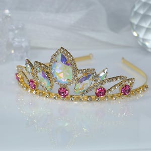 May include: A gold tiara with iridescent and pink rhinestones. The tiara has a delicate design with a pointed center piece.