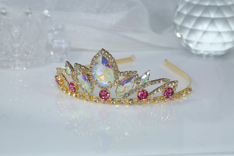 May include: A gold tiara with iridescent and pink rhinestones. The tiara has a delicate design with a pointed center piece.