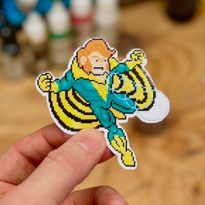 Peut inclure: Un autocollant de personnage pixelisé jaune et bleu avec une cape jaune. Le personnage lève le poing en l'air et a une expression déterminée sur son visage.