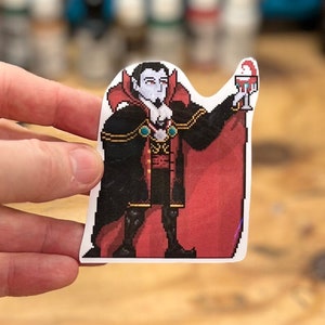 Könnte beinhalten: Ein pixeliger Aufkleber mit einem Vampir, der ein Glas mit roter Flüssigkeit hält. Der Vampir trägt einen schwarzen und roten Umhang und eine goldene Halskette. Der Aufkleber befindet sich auf einer Holzoberfläche.