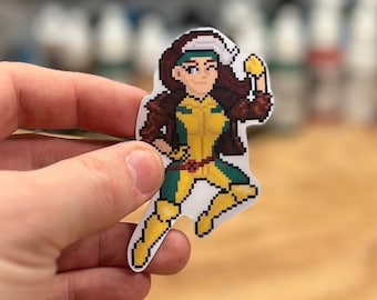 May include: A pixelated sticker of a female superhero character with brown hair, a yellow and green costume, and a white headband. The character is in a fighting pose with one leg raised and a fist clenched.