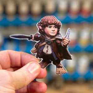 May include: A hand-painted figurine of a hobbit holding a sword and a small bottle. The figurine is made of paper and has a detailed design.