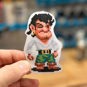 May include: A pixelated sticker of a cartoon pirate character wearing a white shirt, green pants, and a gold belt. The character has black hair and a brown beard.