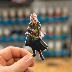 May include: A hand-painted figurine of an elf with long blonde hair, wearing a green and brown tunic, holding a bow and arrow. The elf is standing on a wooden surface.