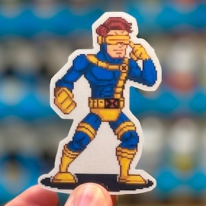 May include: A pixelated sticker of a blue and yellow superhero character with a red headband and yellow goggles. The character is holding their right hand to their face, covering their right eye.
