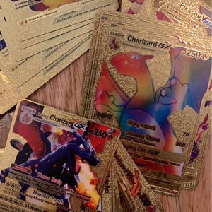 May include: A selection of gold-coloured Pokémon trading cards. The cards feature images of Charizard, Pikachu, and other characters, with text detailing abilities and attacks. The cards have a shiny, metallic finish, and are scattered on a surface.