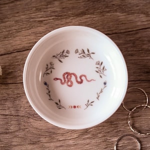 Puede incluir: Un cuenco de cerámica blanco con un diseño de serpiente roja en el centro, rodeado de ilustraciones florales y fases lunares. Tres anillos de plata están junto al cuenco sobre una superficie de madera.