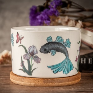 Puede incluir: Un macetero de cerámica blanca con un pez azul y negro, un iris morado y una mariposa rosa. El macetero está sobre un posavasos de madera.