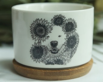 Puede incluir: Maceta de cerámica blanca con una ilustración en blanco y negro de la cara de un perro rodeada de girasoles. La maceta tiene una base de madera.