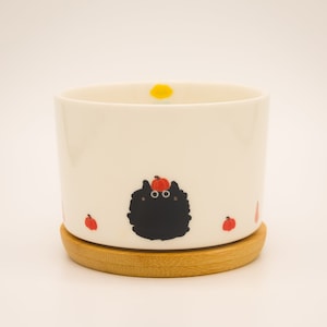 May include: White ceramic planter with a black cat wearing a pumpkin hat and surrounded by small pumpkins. The planter has a wooden base.