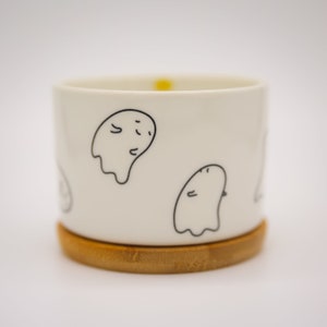 May include: A white ceramic mug with a bamboo coaster. The mug has a black and white ghost design.