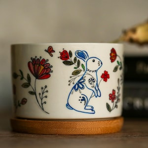 Könnte beinhalten: Weißer Keramik-Pflanztopf mit floralem Design, das ein weißes Kaninchen mit blauen und roten Akzenten zeigt. Der Topf hat einen Holzsockel.